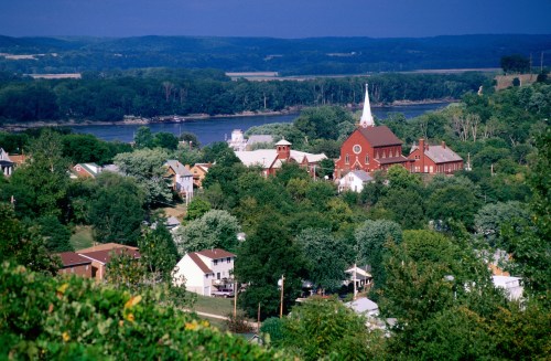 A scenic view of a small town with lush greenery, a river, and a prominent church steeple in the background.