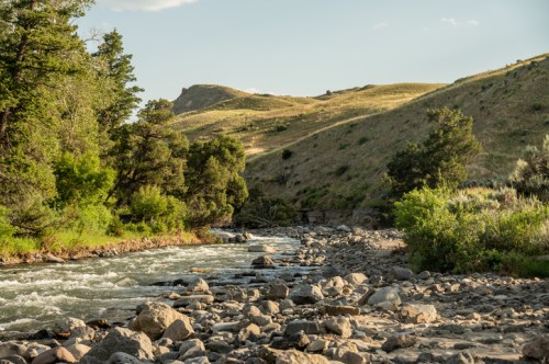 A serene river flows through a rocky landscape, surrounded by lush greenery and rolling hills under a clear sky.
