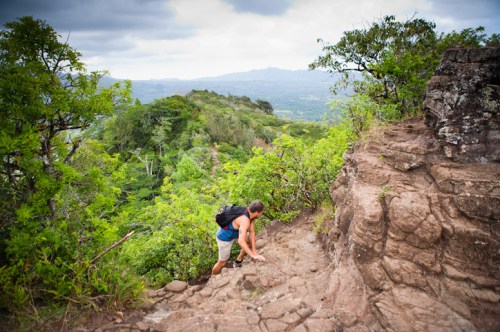 A hiker climbs a rocky trail surrounded by lush greenery and distant hills under a cloudy sky.