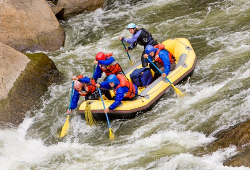 A group of five people in a yellow raft navigating a turbulent river, wearing life jackets and paddling vigorously.