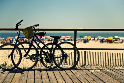 Two bicycles with a basket parked on a wooden boardwalk, overlooking a sunny beach filled with colorful umbrellas.