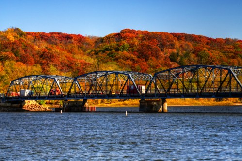 A blue metal bridge spans a river, surrounded by vibrant autumn foliage in shades of orange and yellow.