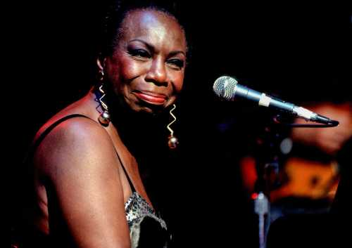 A singer with a microphone, wearing a sparkling dress, gazes emotively at the audience during a performance.