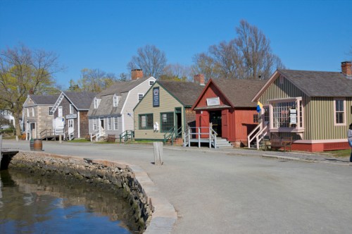 Historic waterfront buildings line a gravel road beside a calm waterway under a clear blue sky.