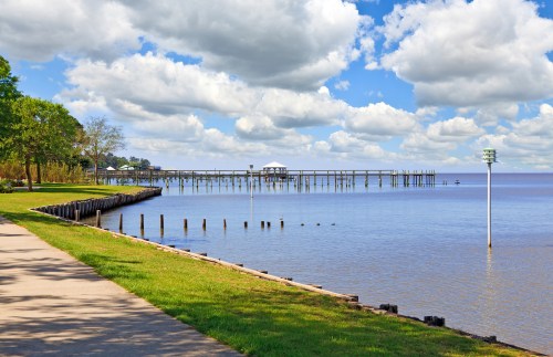 A serene waterfront scene featuring a pier extending into calm waters under a blue sky with fluffy clouds.