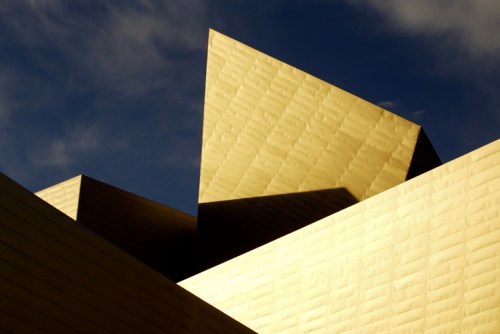Abstract architectural shapes in gold against a blue sky, featuring sharp angles and textured surfaces.