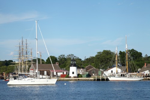 A scenic harbor with sailboats and a lighthouse, surrounded by quaint buildings and greenery under a clear blue sky.