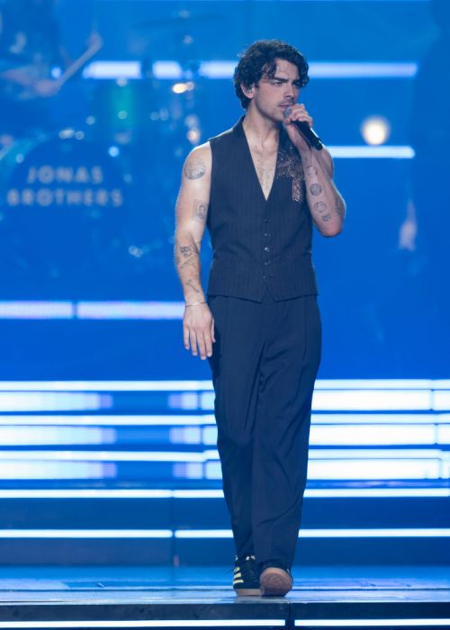 A male singer with tattoos performs on stage, wearing a sleeveless vest and pants, with a band in the background.