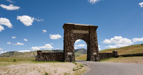 Stone archway with a sign, set against a blue sky and green hills, marking an entrance to a national park.