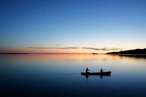 Two people in a canoe paddle across a calm lake at sunset, with a colorful sky reflecting on the water.