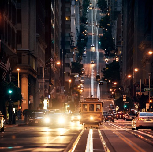 A cable car travels down a steep, illuminated street in a city at dusk, with cars and buildings lining the road.