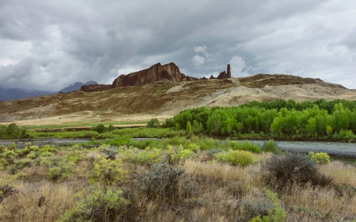 A dramatic landscape featuring rugged mountains, dark clouds, and lush greenery along a riverbank.