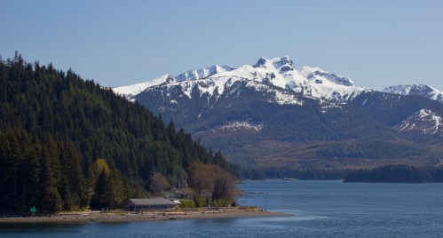 A serene landscape featuring a snowy mountain range, lush green forests, and a calm blue bay under a clear sky.