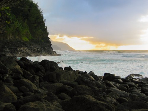 A rocky shoreline with waves crashing against the rocks, under a cloudy sky at sunset.