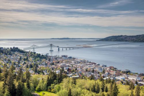 Aerial view of a coastal town with a bridge spanning a river, surrounded by greenery and hills under a cloudy sky.