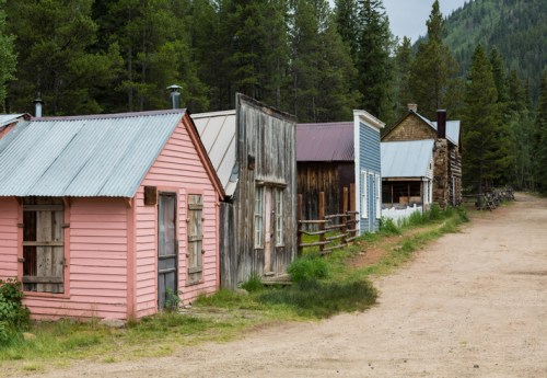 A row of colorful, weathered wooden buildings along a dirt path, surrounded by lush green trees and mountains.