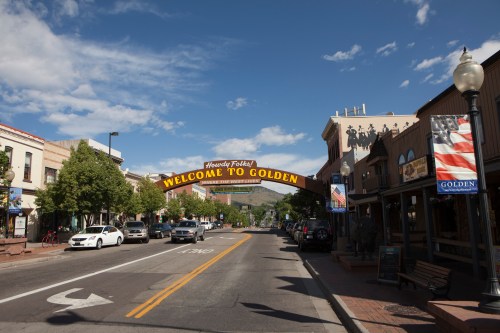 A street view of Golden, Colorado, featuring a welcoming arch and shops under a blue sky with scattered clouds.