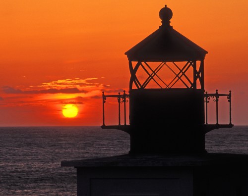 Silhouette of a lighthouse against a vibrant orange sunset over the ocean.