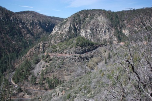 A winding road through mountainous terrain, surrounded by trees and rocky cliffs under a clear blue sky.