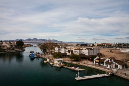 A scenic view of a river with boats, lined by buildings and mountains in the background under a cloudy sky.