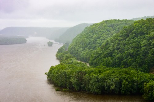 A misty river winding through lush green hills, with dense trees lining the banks under overcast skies.