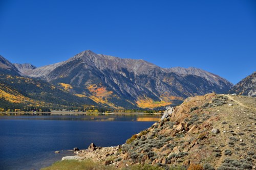 Scenic view of mountains with autumn foliage beside a calm lake under a clear blue sky.