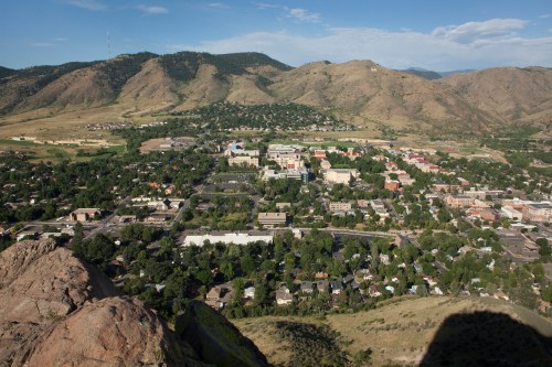 Aerial view of a town surrounded by mountains, featuring green trees and buildings under a clear blue sky.