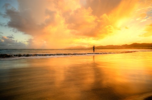 A person stands on a beach at sunset, with vibrant orange and yellow clouds reflecting on the water.