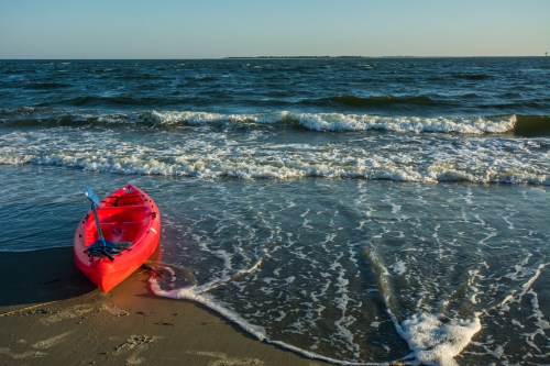 A bright red kayak rests on the shore, with gentle waves lapping at the sand under a clear blue sky.