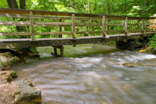 A wooden bridge spans a flowing stream, surrounded by lush green trees and rocky banks.