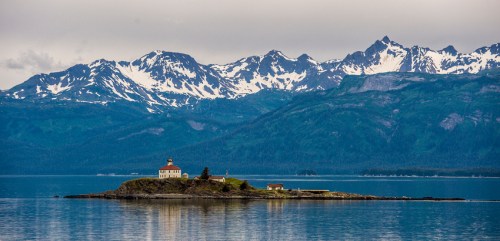 A serene island with a lighthouse surrounded by mountains and calm waters under a cloudy sky.