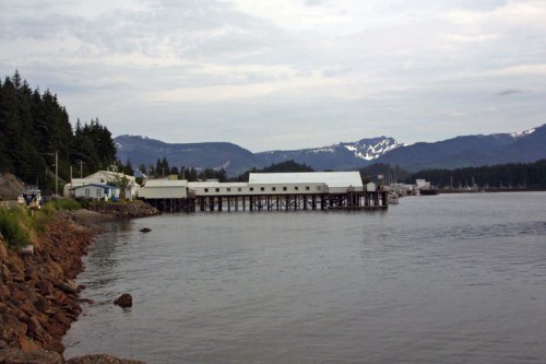 A calm waterfront scene featuring a wooden pier, surrounded by mountains and trees under a cloudy sky.
