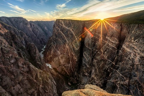 A stunning sunset over a deep canyon, with sun rays illuminating the rocky cliffs and winding river below.