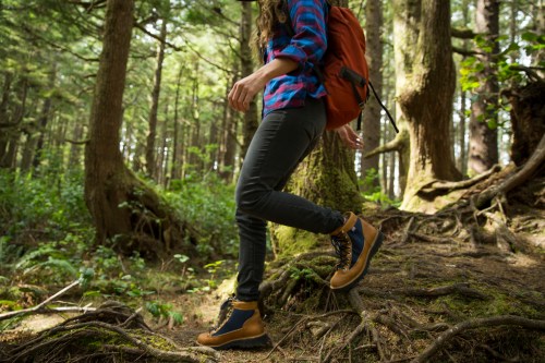 A person wearing hiking boots and a plaid shirt walks along a forest path, surrounded by trees and roots.
