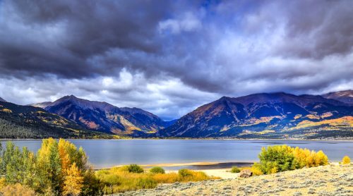 A serene lake surrounded by mountains, with autumn foliage and dramatic clouds in the sky.