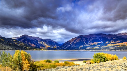 A serene lake surrounded by mountains, with autumn foliage and dramatic clouds in the sky.