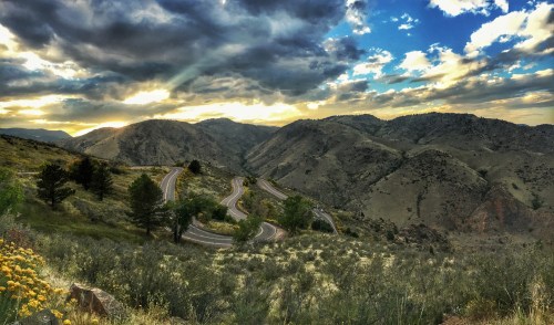 Winding road through mountainous terrain under a dramatic sky at sunset, with lush greenery and wildflowers in the foreground.