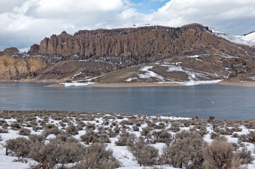 A serene landscape featuring a frozen lake, snow-covered hills, and rugged cliffs under a cloudy sky.