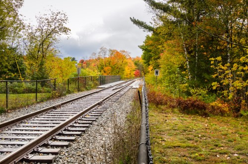 A view of train tracks stretching into the distance, surrounded by vibrant autumn foliage and a cloudy sky.