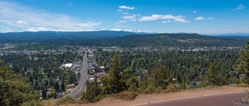 A panoramic view of a town surrounded by trees and mountains under a clear blue sky.