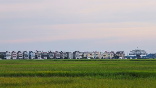 A row of colorful houses along a marshy area under a pastel sky, with a bridge visible in the distance.