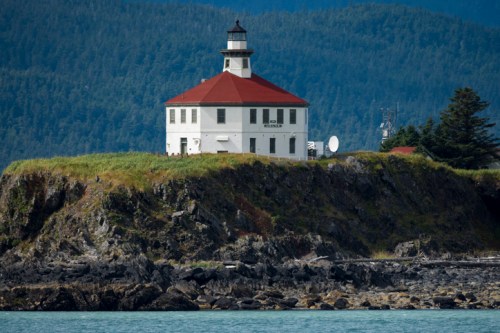 A white lighthouse with a red roof sits on a rocky shore, surrounded by green hills and blue water.
