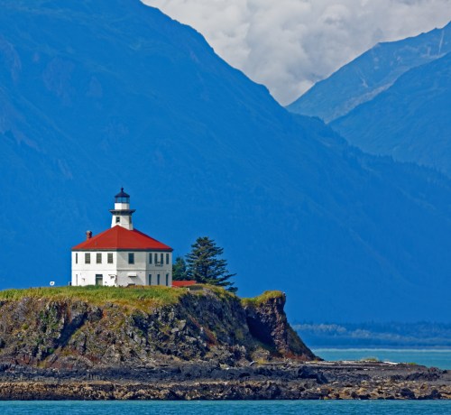 A white lighthouse with a red roof sits on a rocky island, surrounded by mountains and a calm blue sea.