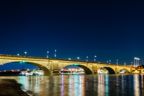 A lit bridge spans a river at night, with boats docked nearby and stars visible in the dark sky.