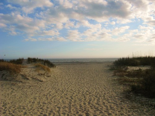 A sandy path leads to the ocean, framed by grass and dunes under a cloudy sky.