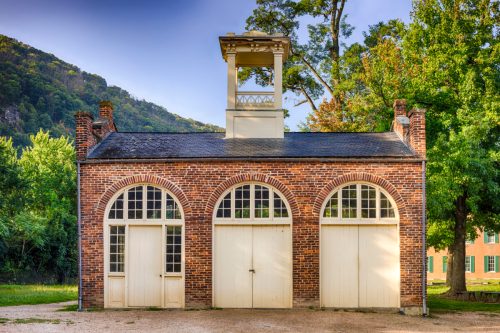 A brick building with arched windows and a bell tower, surrounded by trees and mountains in the background.