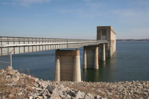 A concrete bridge extends over calm water, supported by large pillars, with rocky terrain in the foreground.