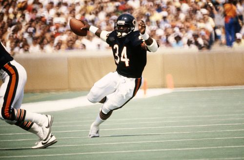A football player in a dark jersey throws a pass on the field during a game, with a crowd in the background.