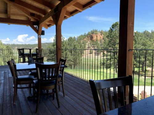 A wooden deck with tables and chairs overlooks a green landscape and trees under a blue sky.