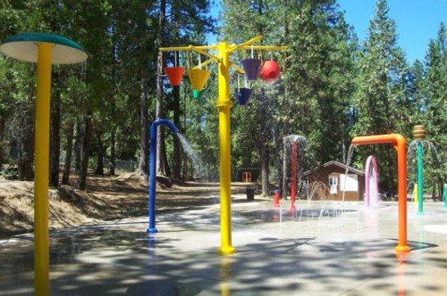 Colorful water play area with various spray features surrounded by trees and a wooden structure in the background.